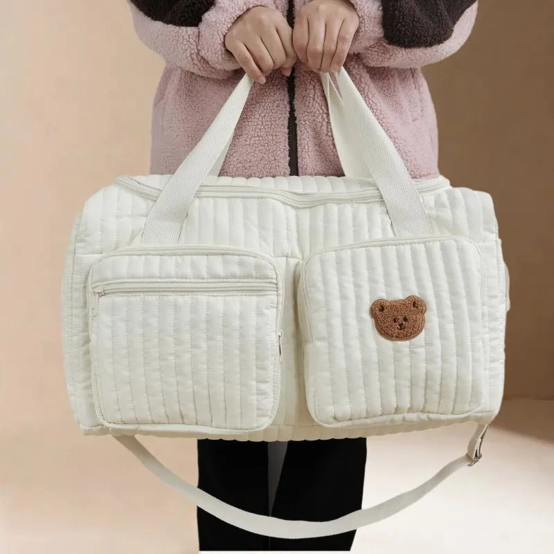 Bolsa maternidade acolchoada off-white com patch de ursinho marrom, segurada de frente.