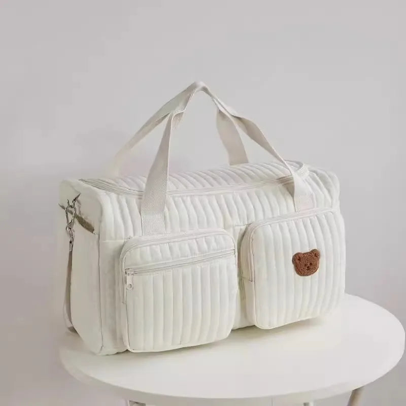 Bolsa maternidade branca acolchoada com dois bolsos e ursinho bordado sobre mesa redonda.