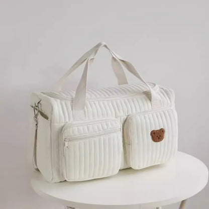 Bolsa maternidade branca acolchoada com dois bolsos e ursinho bordado sobre mesa redonda.