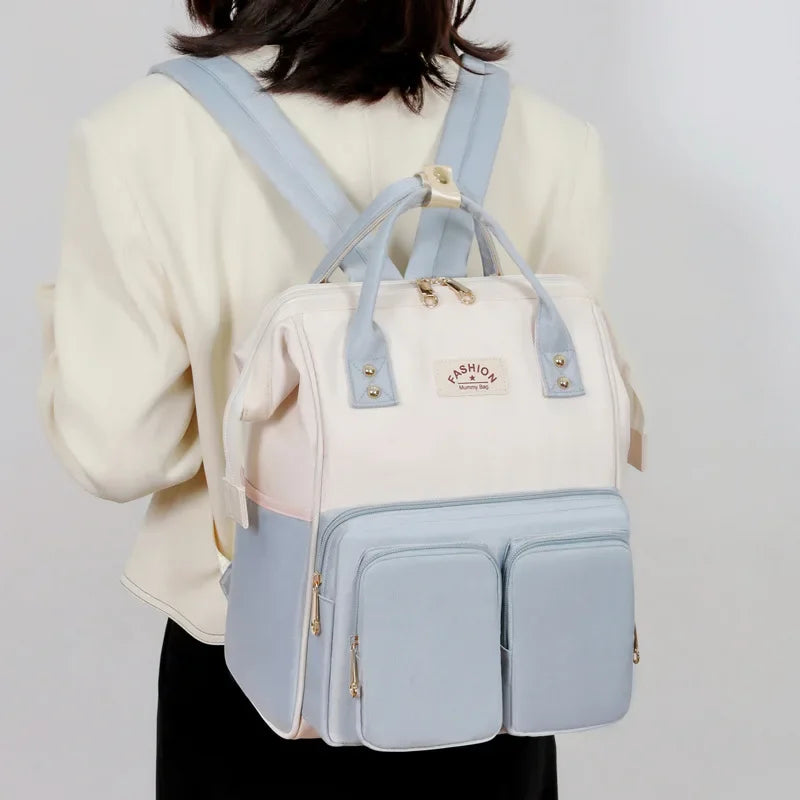 **Alt Text:** Mochila maternidade multifuncional azul pastel e creme, vista de costas em uma modelo.