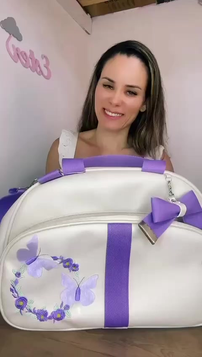 Mala de maternidade branca arredondada com alças, laço e bordado de borboletas lilás.