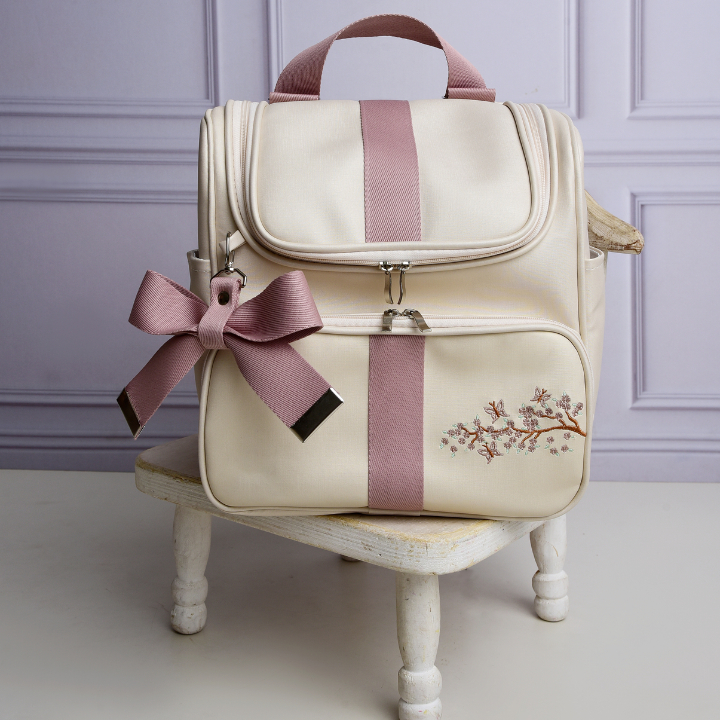Mochila de maternidade bege com faixas rosa, laço e bordado floral, vista de frente.