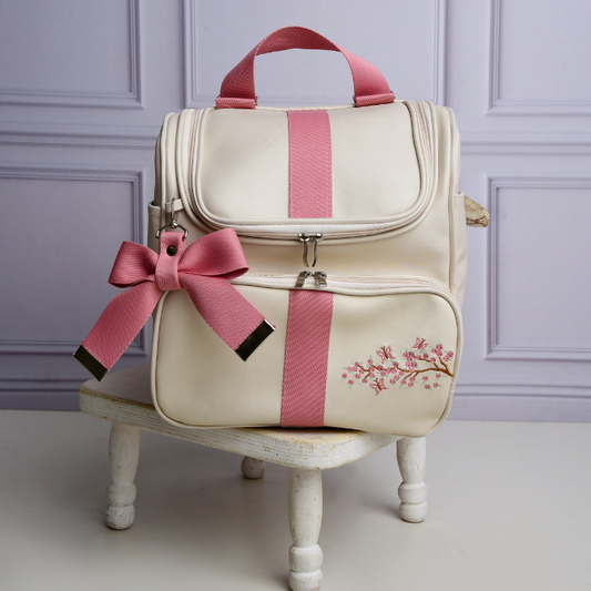 Mochila de maternidade bege e rosa com bordado floral, apoiada em banqueta branca.