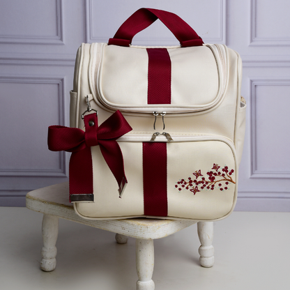 Mochila de maternidade bege com detalhes bordô e bordado floral, sobre pequeno banco branco.
