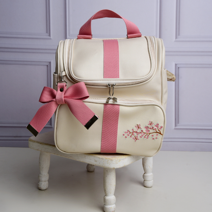 Mochila de maternidade creme com detalhes rosa, laço decorativo e bordado floral, vista frontal.