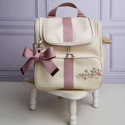 Mochila de maternidade bege com listra rosa, bordado de flores e laço decorativo.