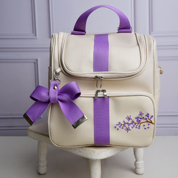 Mochila bege estruturada de frente com listras roxas, bordado floral e laço decorativo.