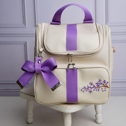 Mochila bege estruturada de frente com listras roxas, bordado floral e laço decorativo.