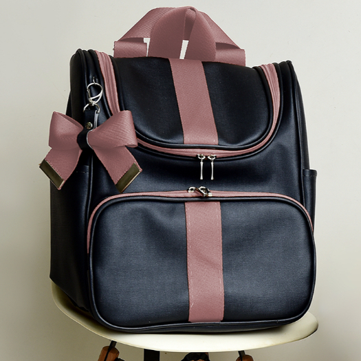 Mochila preta com detalhes e laço rosa, vista de frente sobre banqueta branca.