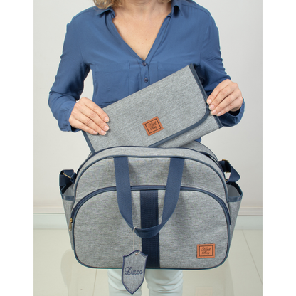 Bolsa de maternidade cinza com detalhes azuis, trocador e chaveiro personalizado em vista frontal.