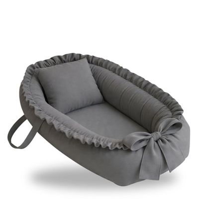 Ninho de bebê oval cinza com travesseiro, bordas franzidas, laço frontal e alça lateral.