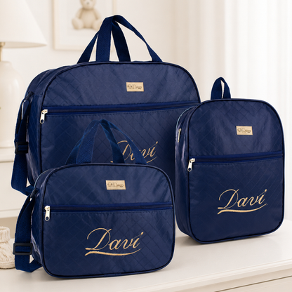 Três bolsas de maternidade azul-marinho personalizadas com bordado dourado Davi sobre mesa.