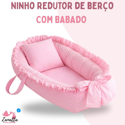 Ninho redutor de berço rosa oval, com babados, laço e travesseiro, em ângulo inclinado.
