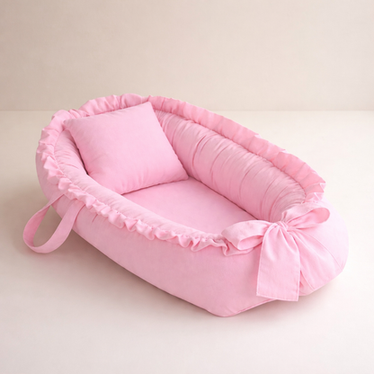 Ninho de bebê rosa com travesseiro, babados e laço, fotografado em ângulo diagonal.