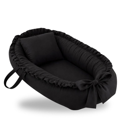 Ninho de bebê oval preto com travesseiro e laço frontal, visto em ângulo inclinado.