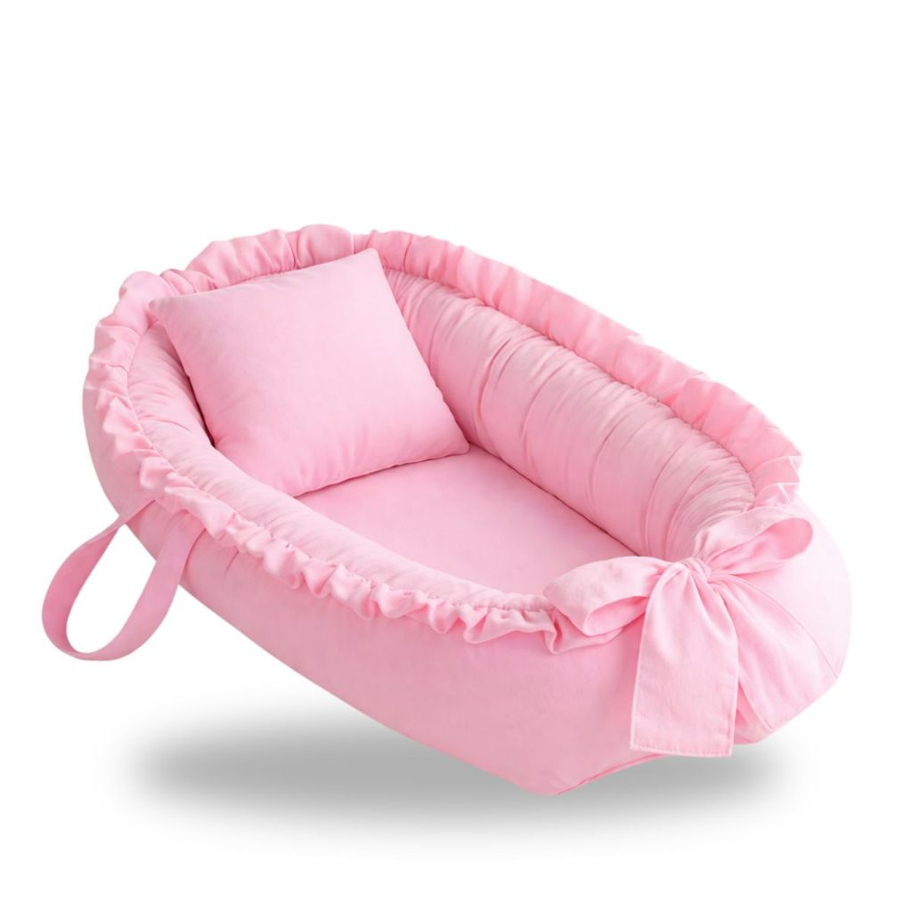 Ninho de bebê oval rosa claro com travesseiro e laço, exibido em ângulo diagonal.